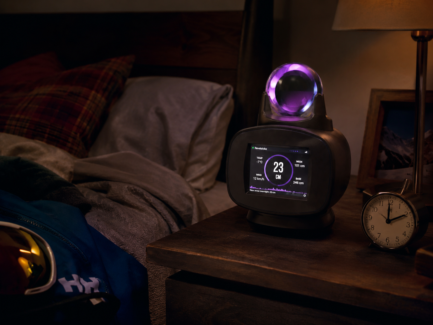 The Powder Oracle glowing on a bedside table at night, its screen reading 23 cm of fresh snow overnight beside a ski jacket and goggles